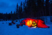Snöskor utanför upplyst tält med sovande hundspann utanför. Explore Sarek Nationalpark.